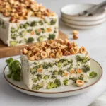 Broccoli, hazelnut terrine slice on white plate. Creamy base with broccoli florets and whole nuts on top.