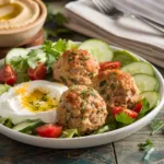 Mediterranean Baked Turkey Meatballs served with hummus, a side of Mediterranean salad, and a drizzle of yogurt sauce, garnished with parsley and lemon zest on a white plate.