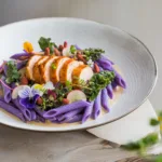 A vibrant plate of purple butterfly pea pasta with golden turmeric-crusted chicken, garnished with edible flowers, microgreens, and watermelon radish slices.